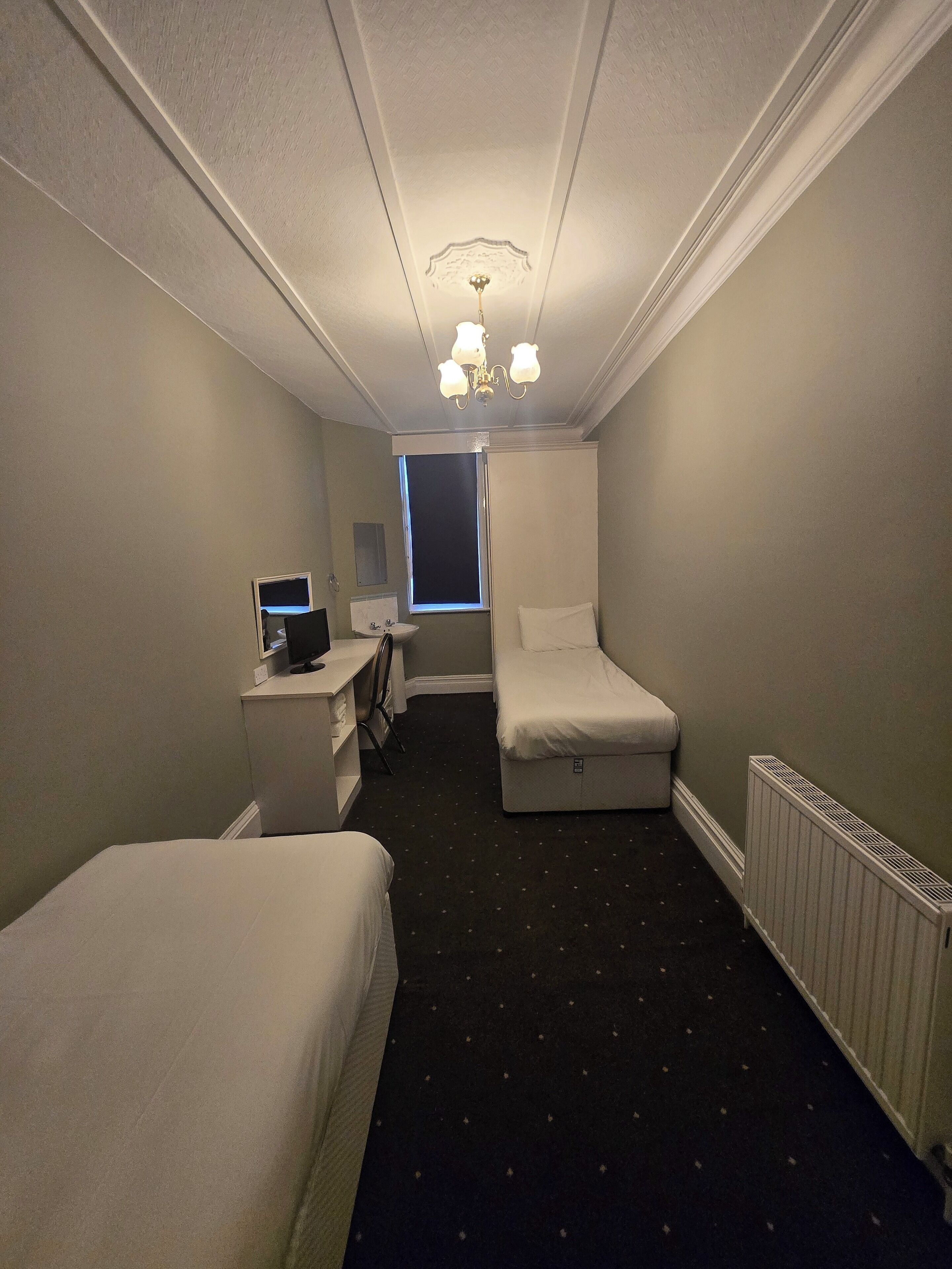 A hotel room with two single beds, a desk with a computer, a window, and a radiator.