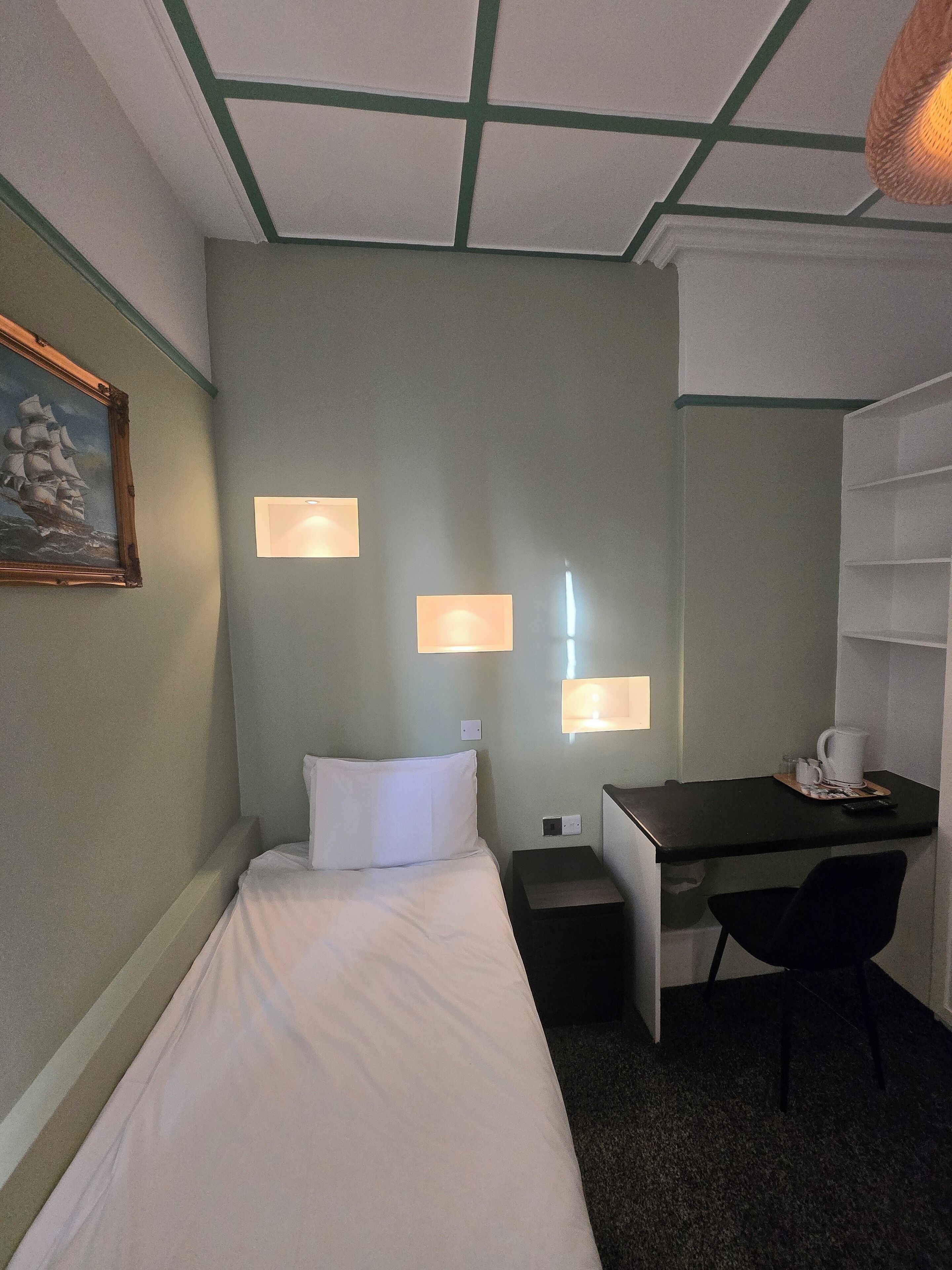 A small hotel room with a bed, desk, chair, and two wall-mounted lamps.