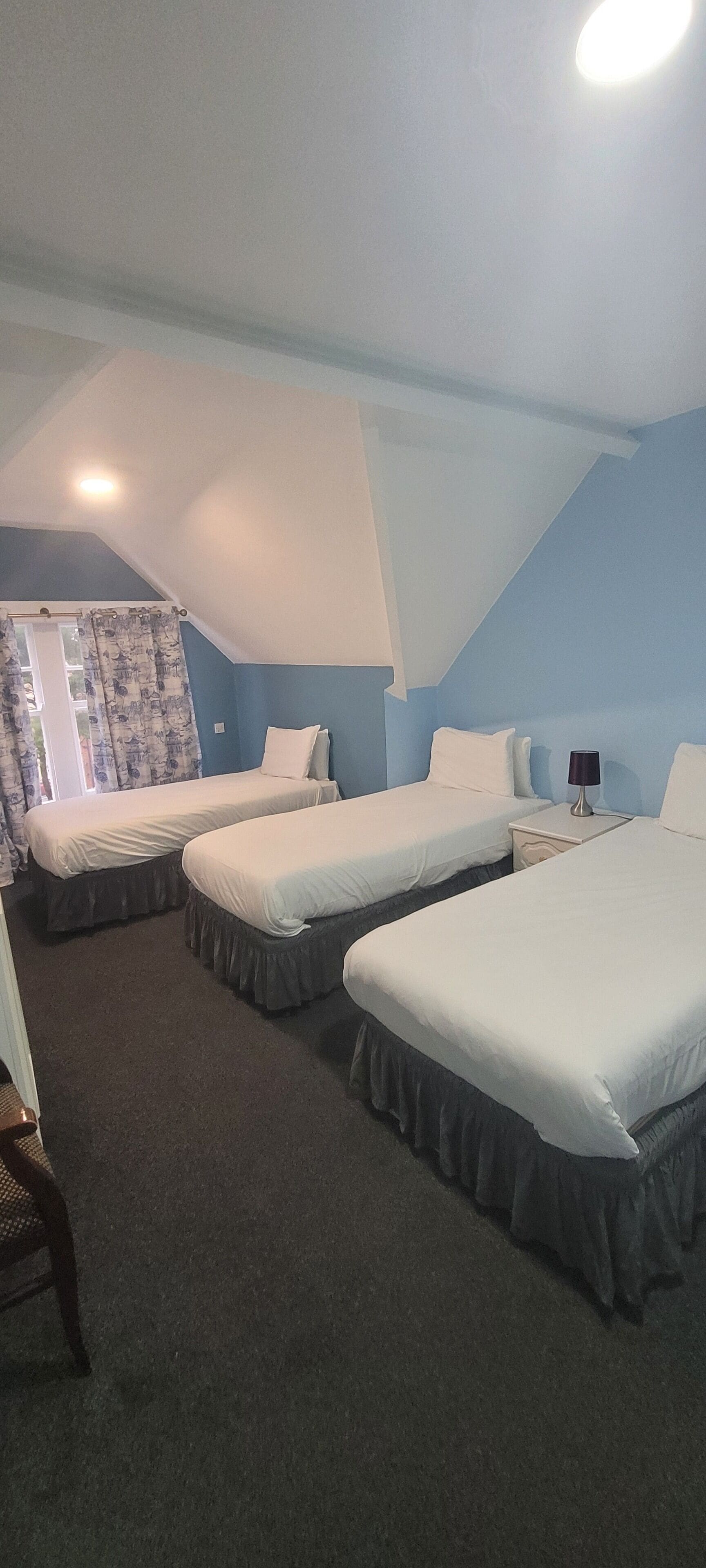 A room with three single beds, a small table with a lamp, and a window with curtains.
