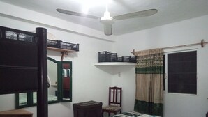 A bedroom with a bed, a chair, a small table, a trash bin, a ceiling fan, a window with curtains, and a shelf with items. - Divers Hostel Cozumel (Cozumel)