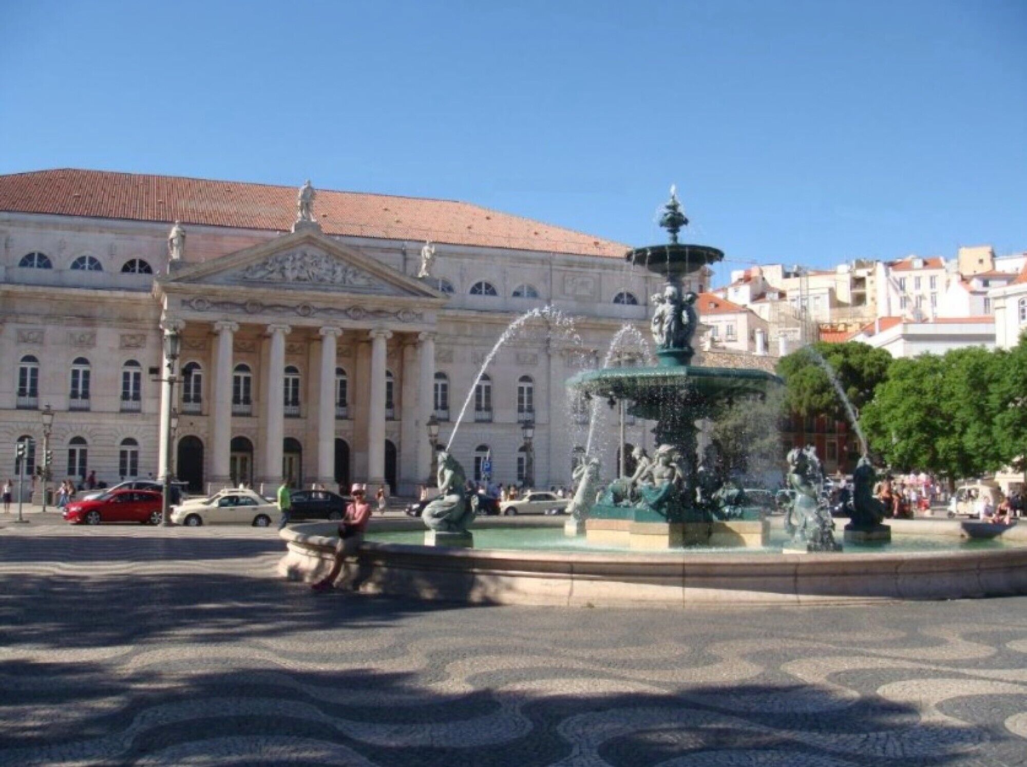 A public square with a fountain, a large classical building with columns, and a clear blue sky.