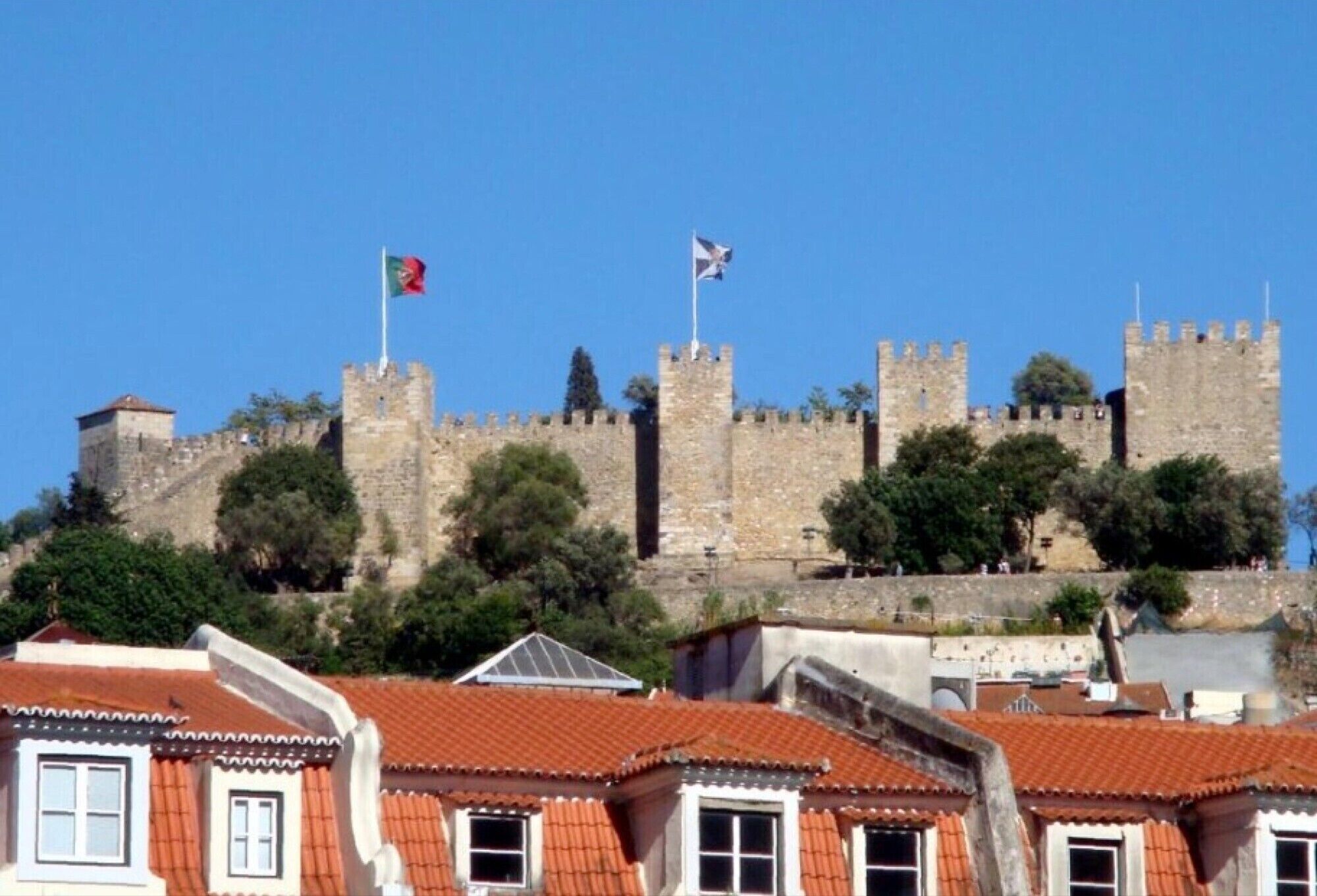 A historic castle with stone walls and flags, surrounded by buildings with red-tiled roofs.