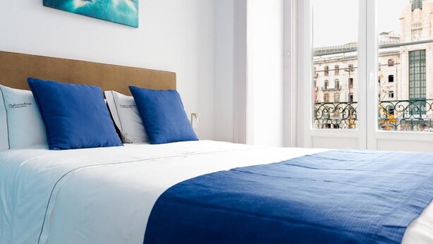 A bed with a white and blue bedspread, two blue pillows, and a view of a cityscape through a window. - 'Inn Chiado - Double Room 2' with Balcony, Wi-Fi and Air Conditioning (Lisboa)