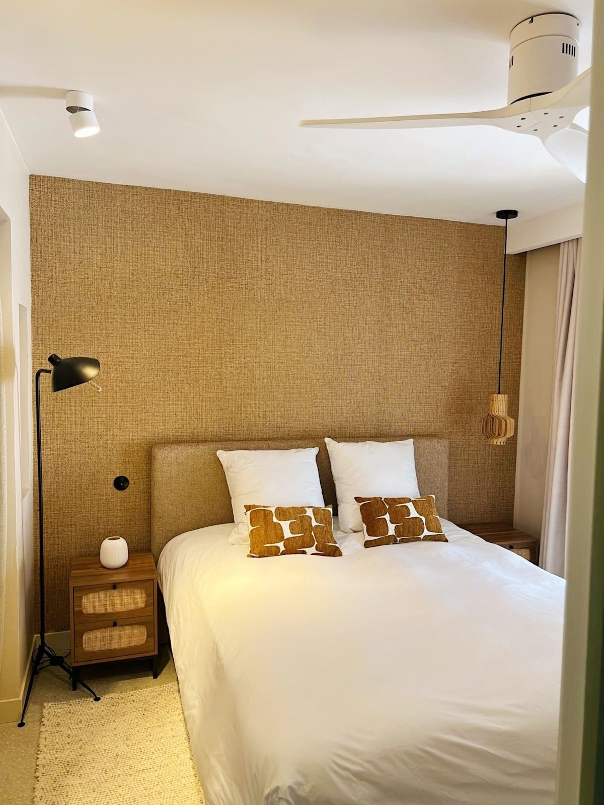 A hotel room with a large bed, a nightstand, a lamp, a ceiling fan, and a textured wall.