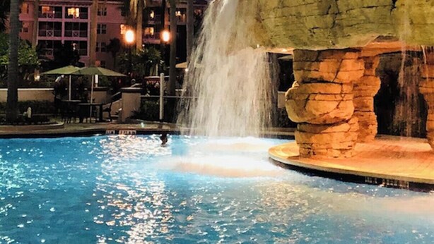 Ein Pool mit Wasserfall, umgeben von Palmen und einem mehrstöckigen Gebäude im Hintergrund.