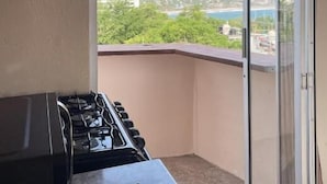 A balcony with a gas stove, a sliding glass door, and a view of a coastal area with greenery and buildings. - Casa Laani (San Pedro Mixtepec)
