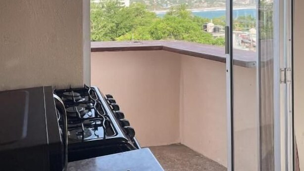 Un balcon équipé d’une cuisinière à gaz, d’une porte-fenêtre coulissante et offrant une vue sur une zone côtière verdoyante, avec des bâtiments.