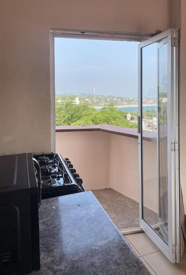 A balcony with a gas stove, a sliding glass door, and a view of a coastal area with greenery and buildings. - Casa Laani (San Pedro Mixtepec)