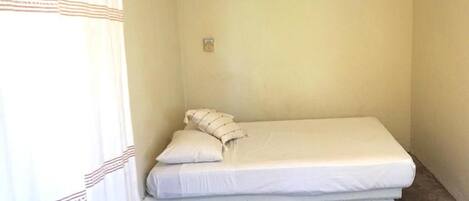 Una habitación pequeña con cama individual, colchón y almohadas sencillos, pared de color claro y una ventana con cortina.