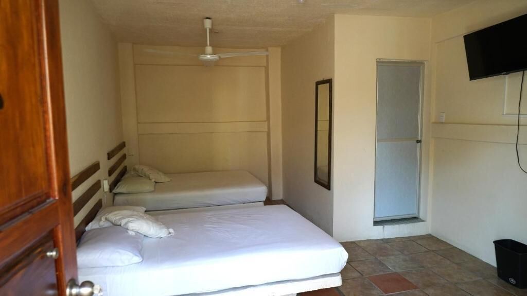 Una habitación pequeña y limpia con dos camas individuales, un ventilador de techo, una ventana y un televisor montado en la pared.