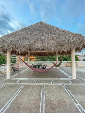 A thatched-roof gazebo with a hammock and chairs, located on a rooftop with a view of buildings and greenery. - Casa Laani (San Pedro Mixtepec)