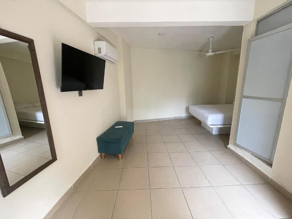 A room with a flat-screen TV mounted on the wall, a small teal ottoman, a bed with a white mattress, and a sliding glass door.