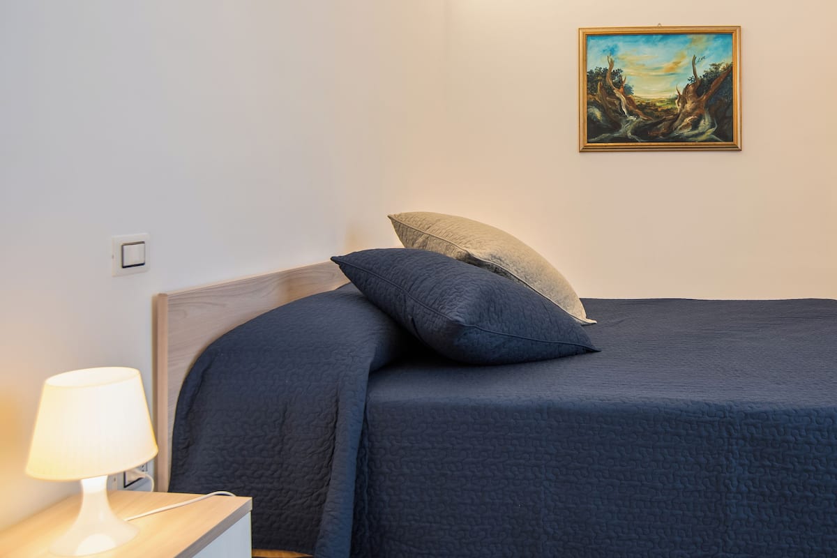 A neatly made bed with a dark blue bedspread and pillows, a bedside lamp, and a framed painting on the wall.