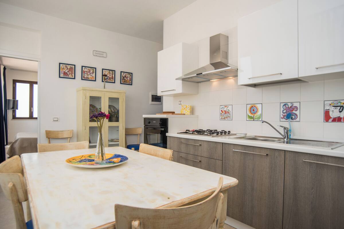 A modern kitchen with white cabinets, a gas stove, and a sink. There is a dining table with chairs and a vase with flowers.
