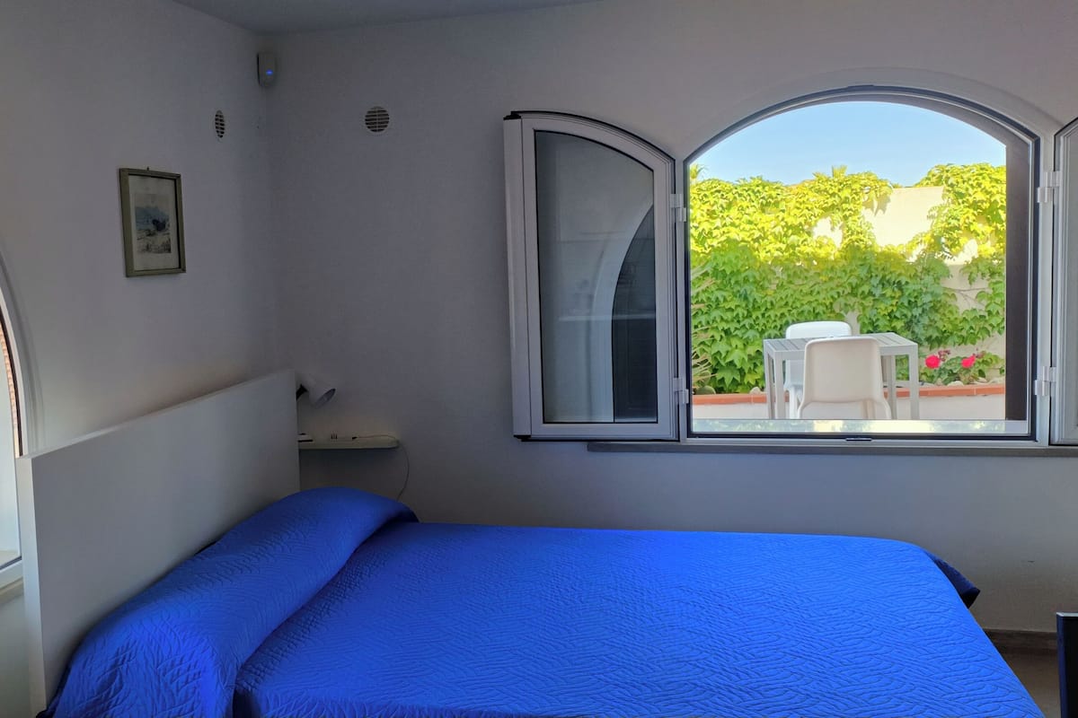 A bedroom with a bed, two windows, a bedside table, and a view of a balcony with outdoor furniture.