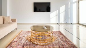 A modern living room with a beige sofa, a round glass coffee table with a gold frame, and a patterned rug. A flat-screen TV is mounted on the wall.