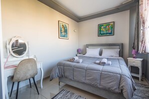 A bedroom with a bed, a desk with a chair, a mirror, and two paintings on the wall. - Nancy View Apartment - Right Next to the Airport (Markopoulo Mesogaias)