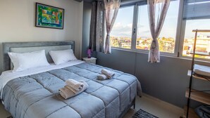 Une chambre à coucher avec un lit, une table de chevet, une fenêtre avec des rideaux, une vue sur des immeubles et un tableau accroché au mur.