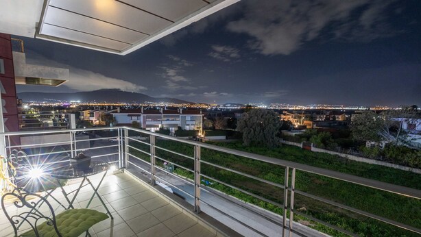 Un balcon avec un fauteuil à coussins verts, une petite table sur laquelle se trouve un bol, et une balustrade d’où l’on peut admirer la vue sur la ville la nuit.