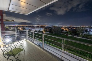 A balcony with a green cushioned chair, a small table with a bowl, and a railing overlooking a cityscape at night. - Nancy View Apartment - Right Next to the Airport (Markopoulo Mesogaias)