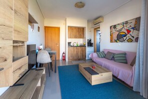 A modern living room with a wooden cabinet, a blue rug, a wooden coffee table, a pink sofa, and a colorful abstract painting on the wall. - Nancy View Apartment - Right Next to the Airport (Markopoulo Mesogaias)
