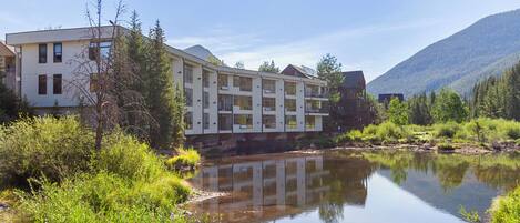 A multi-story hotel with a river in front, surrounded by greenery and mountains.