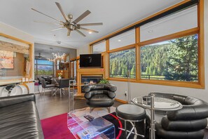 A modern living room with a fireplace, leather furniture, a television, and a large window with a forest view. - Condo in River Run Village! Fireplace! 5 Minute Walk to the Ski Lift! (Keystone)