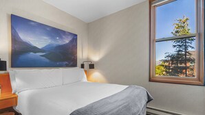 Ein Schlafzimmer mit einem Bett, einem Nachttisch, einer Wandlampe, einem Gemälde und einem Fenster mit Blick auf Bäume und ein Gebäude.
