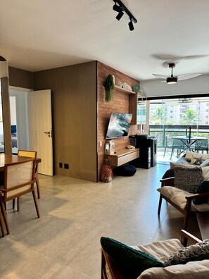 A modern living room with a TV mounted on a brick wall, a dining area with a wooden table and chairs, and a balcony with outdoor furniture. - Beachfront Apartment Set Back from the Setback (Bertioga)