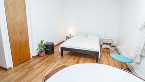 Et soveværelse med trægulv, seng med hvidt sengetøj, et natbord, en krukke med plante og en blå bænk.