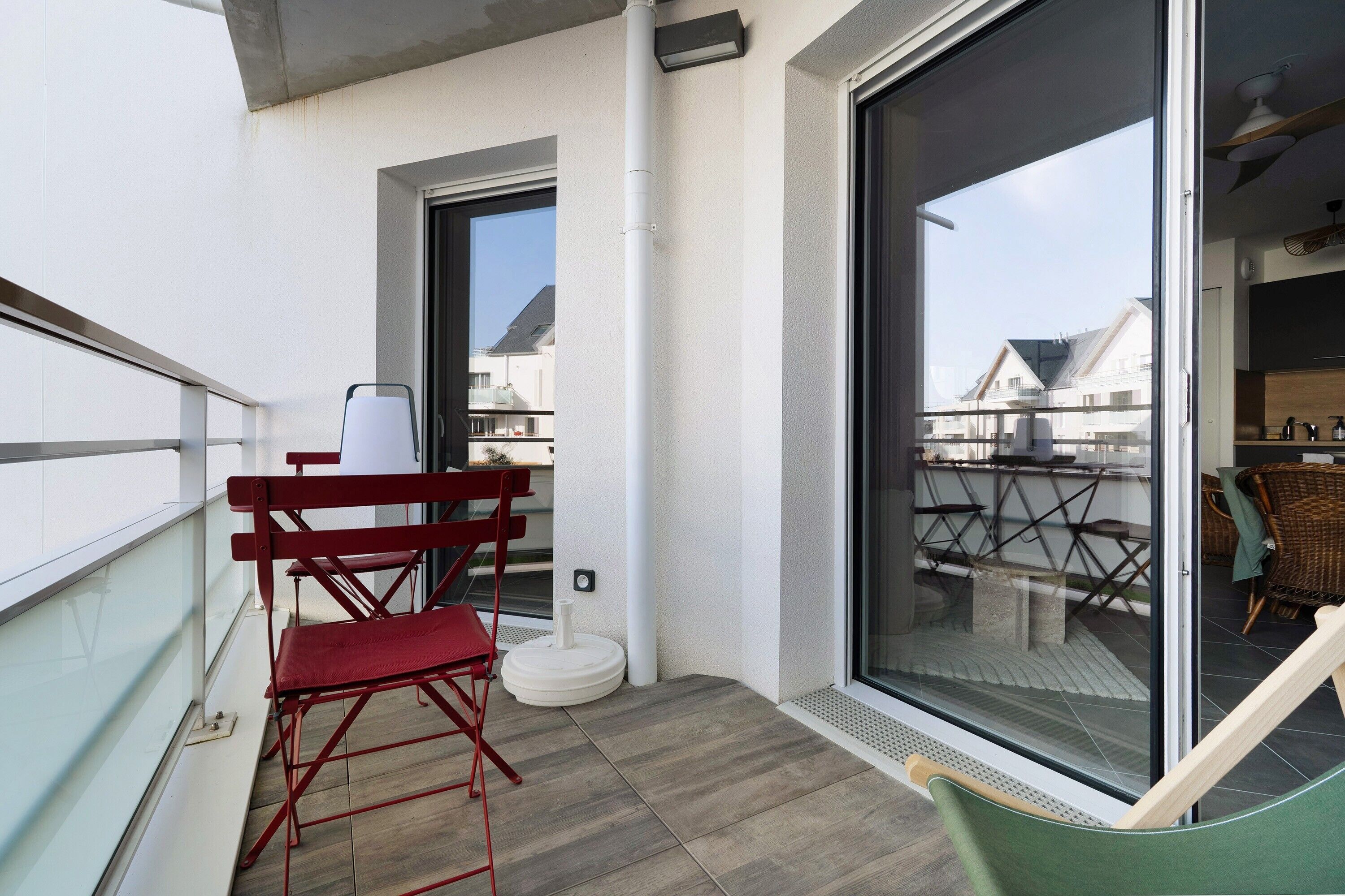 Un balcon avec une chaise pliante rouge, une chaise verte et une porte vitrée donnant sur un espace extérieur aménagé pour s’asseoir.