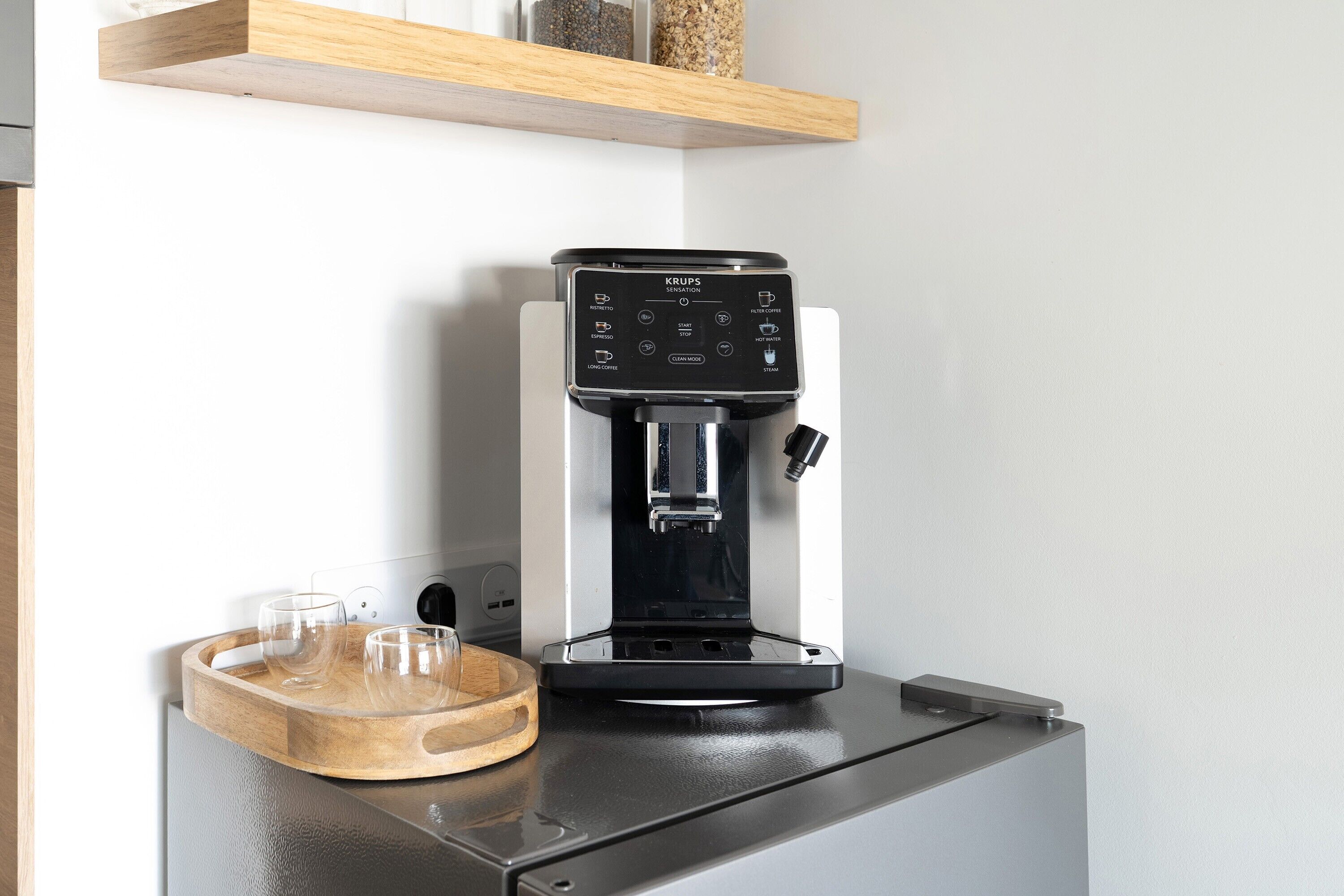 Une machine à café moderne posée sur un plan de travail de cuisine, avec un plateau en bois et des verres.