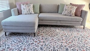 Un canapé d’angle gris avec des coussins à motifs floraux et à rayures, un pouf assorti et un tapis à motifs floraux.