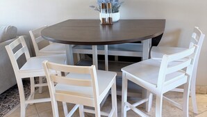 Une table de salle à manger ronde, entourée de quatre chaises blanches, un vase avec des fleurs et, au fond, un canapé gris.