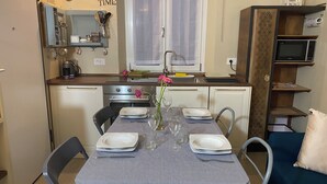 Una cocina compacta con zona de comedor. Incluye cocina, horno, fregadero y un refrigerador pequeño. Hay una mesa para dos personas con platos, vasos y un florero con flores.
