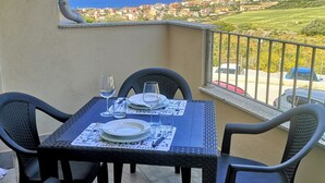 A balcony with a table set for two, chairs, and a view of the sea and town. - Sea View Residence with Pool (Lu Bagnu)