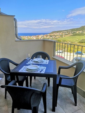A balcony with a table set for two, chairs, and a view of the sea and town. - Sea View Residence with Pool (Lu Bagnu)