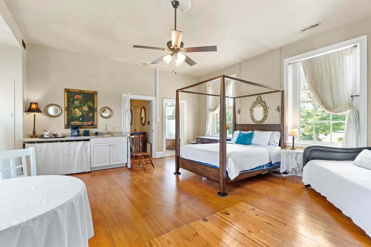 Un dormitorio con una cama con dosel, un escritorio con silla, una mesa de comedor y un ventanal con cortinas.