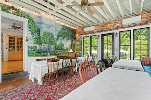 A room with a mural of trees and a house, a dining area with a table and chairs, and large windows with a view of greenery.