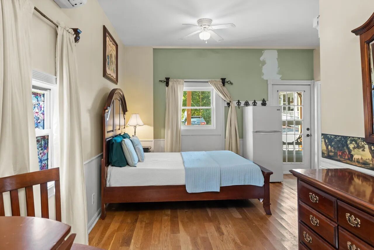 Une chambre à coucher avec un lit en bois, une table de chevet, une commode, un ventilateur de plafond et une fenêtre avec des rideaux.