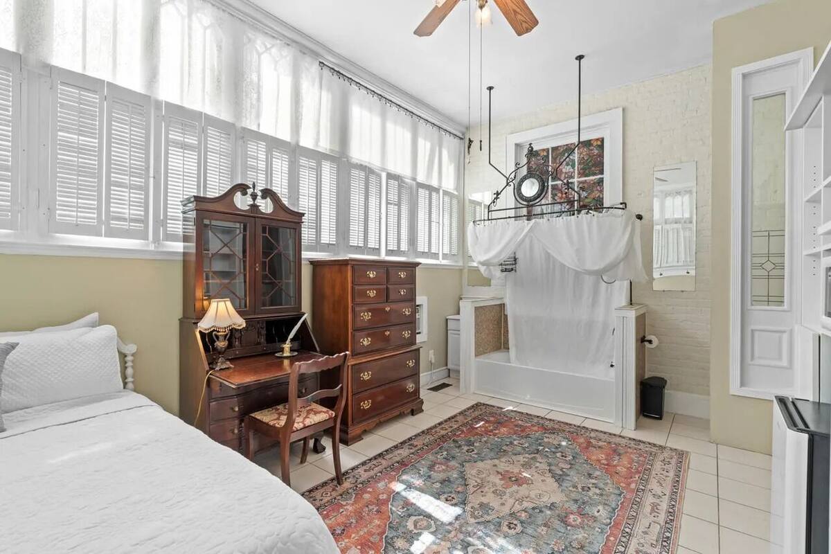 Un dormitorio con cama, cómoda de madera, escritorio, silla y un ventanal con contraventanas.