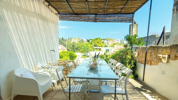 Un patio exterior con una mesa de vidrio y sillas blancas, techo de paja y vista de edificios al fondo.