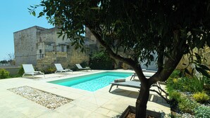 Una piscina con sillones de descanso, un árbol y un edificio de piedra al fondo.