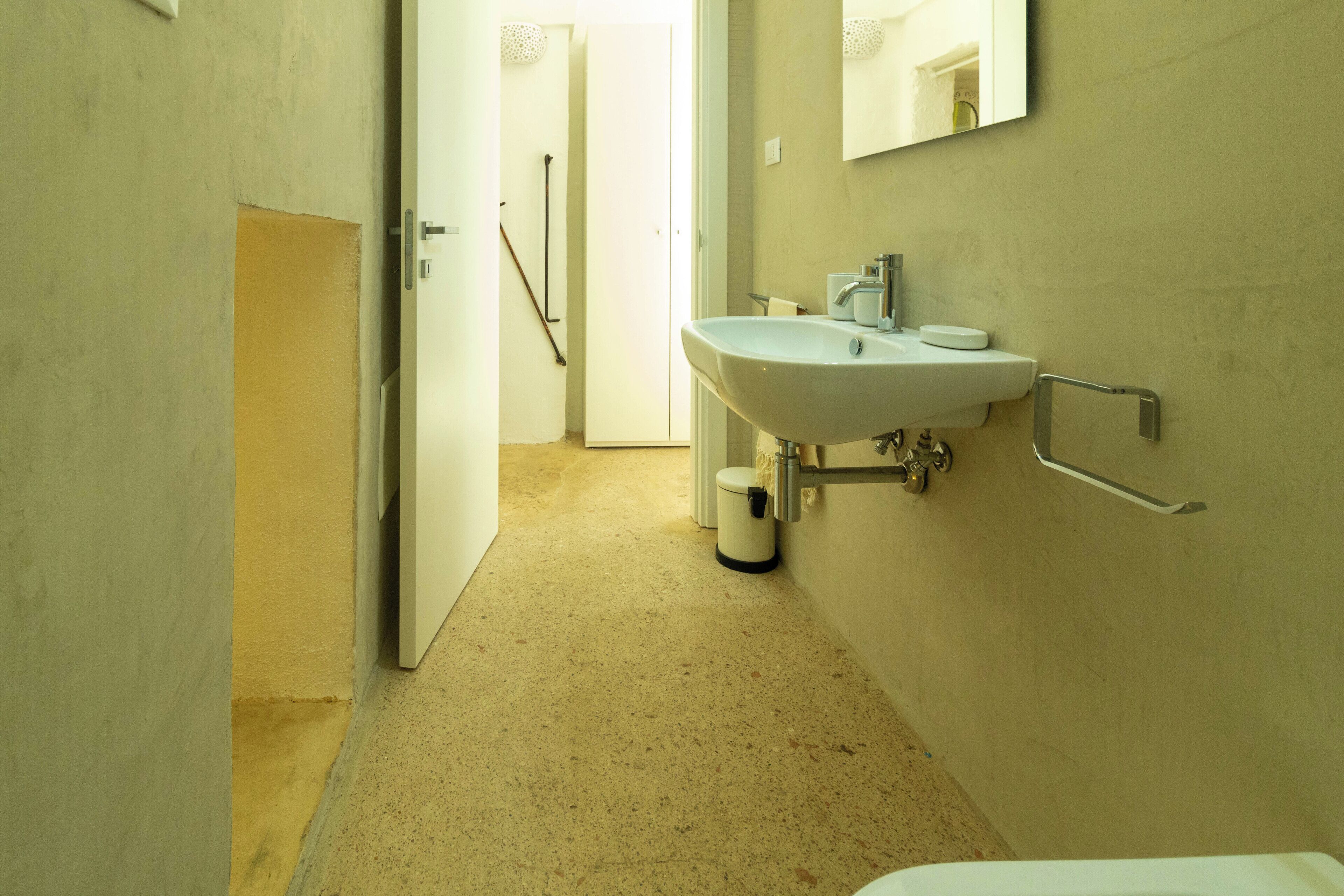 A narrow hallway with a white door, a beige tiled floor, and a white sink with a mirror above it.