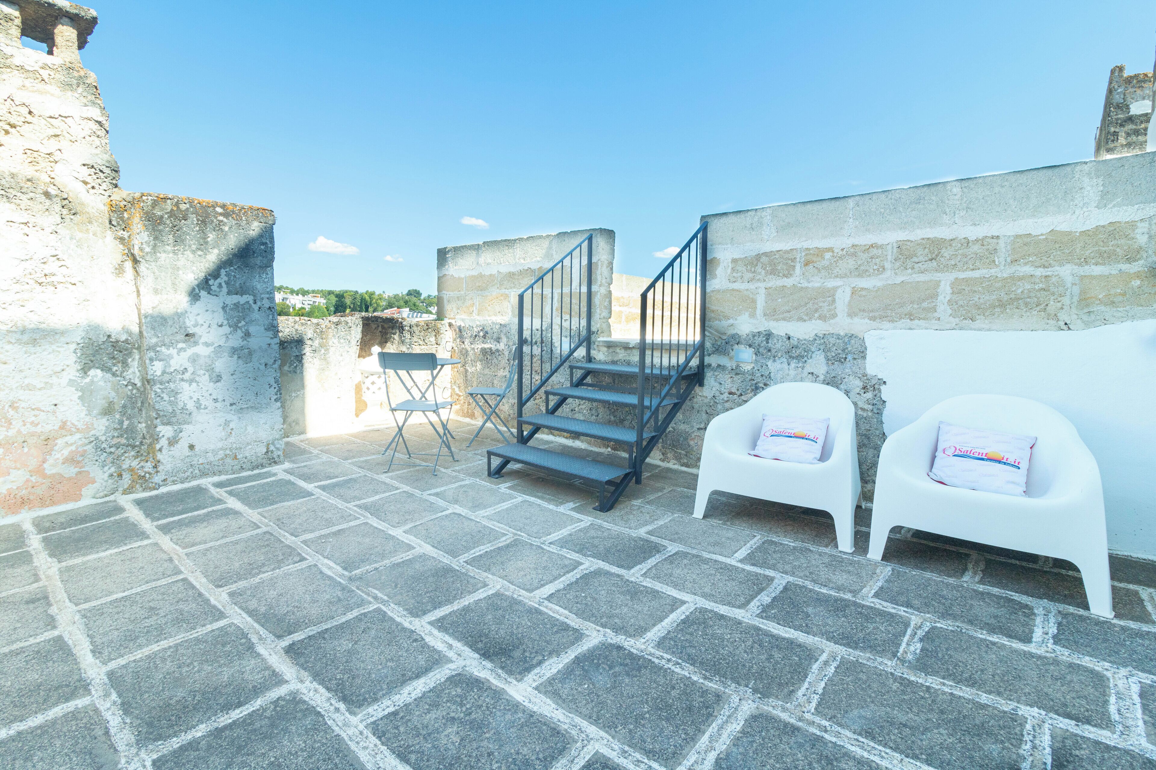 A courtyard with stone flooring, a metal staircase, and two white plastic chairs with cushions.