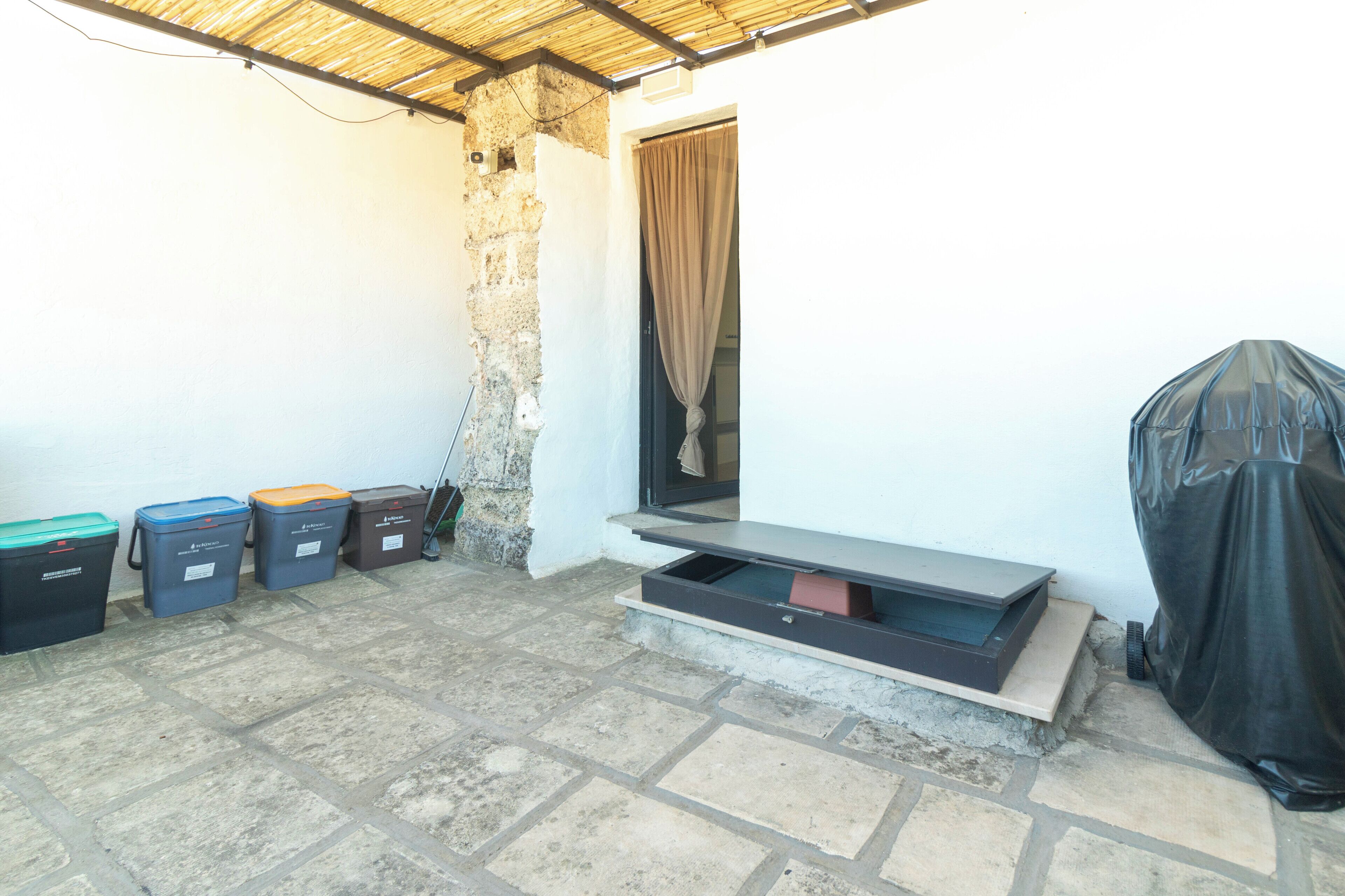 A small outdoor area with a stone floor, a bench, and trash bins.