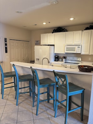 A kitchen with white cabinets, a white refrigerator, a microwave, a coffee maker, and three blue chairs.