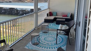 A covered balcony with a view of a lake, a seating area with cushions, and a patterned rug. - Spacious waterfront 3-bedroom condo in pleasant Sunrise Beach with pool access (Sunrise Beach)