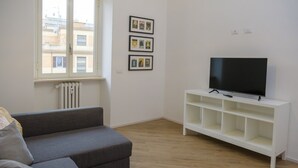 A living room with a grey sofa, a white TV stand, and a window with a view of buildings. - Rome Urban Retreat - ChiacchioProperty (Rome)