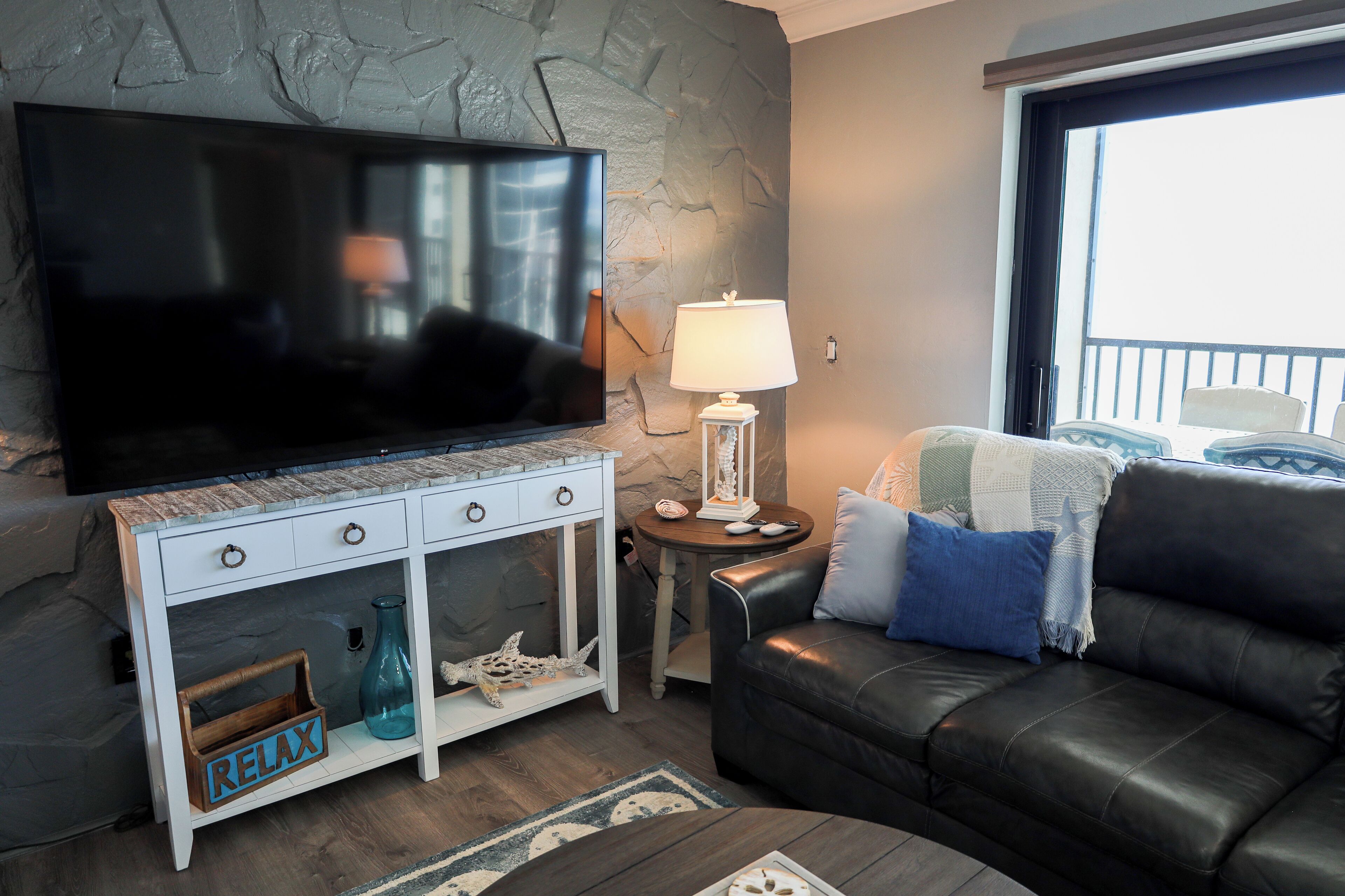 A living room with a flat-screen TV, a white console table with drawers, a black leather sofa, and a lamp.